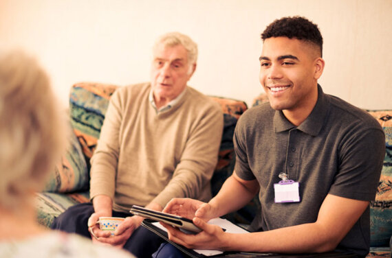 Young social worker sitting on the couch with an older man, with tablet in hand