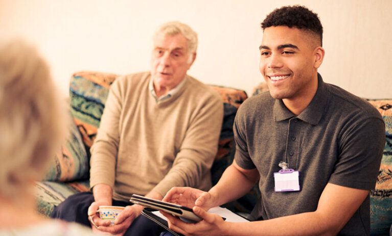 Young social worker sitting on the couch with an older man, with tablet in hand