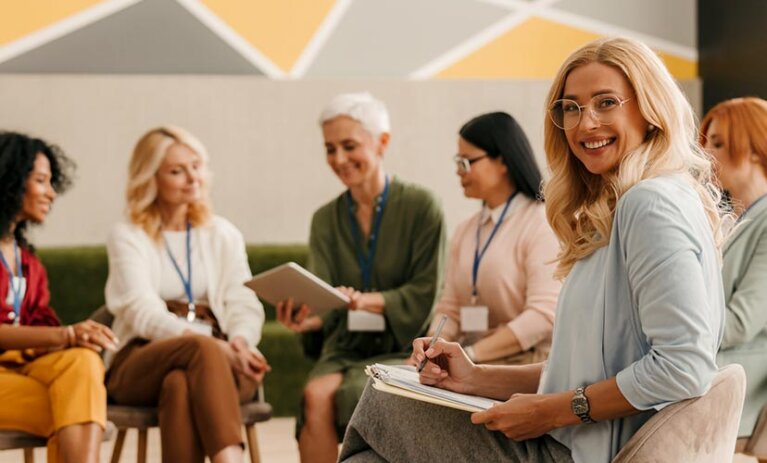 A group of woman sitting on chairs in a semicircle, with a young smiling woman turning to face camera in the foreground