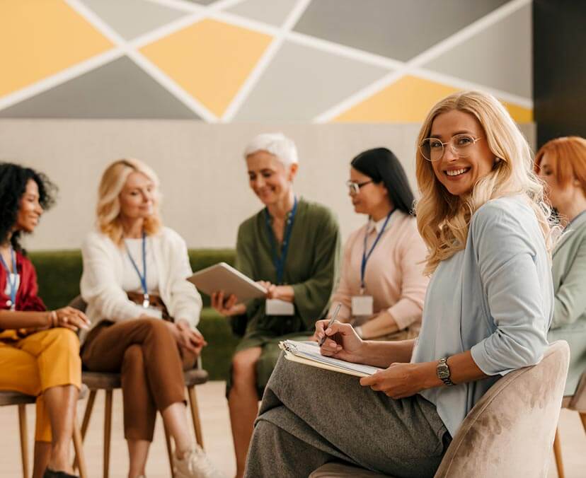 Coaching A group of woman sitting on chairs in a semicircle, with a young smiling woman turning to face camera in the foreground