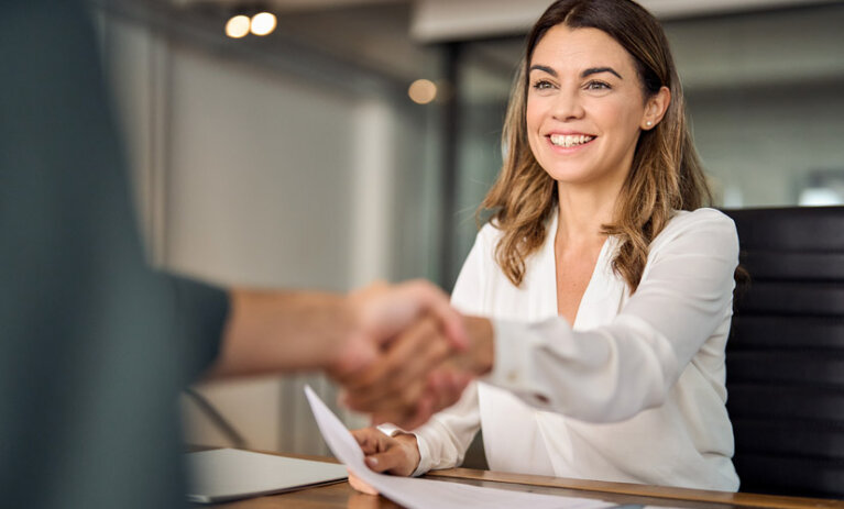 Young smiling woman sitting, shaking someone's hand over a work desk