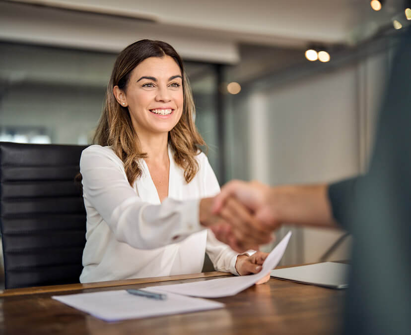 Young smiling woman sitting, shaking someone's hand over a work desk (left of screen)