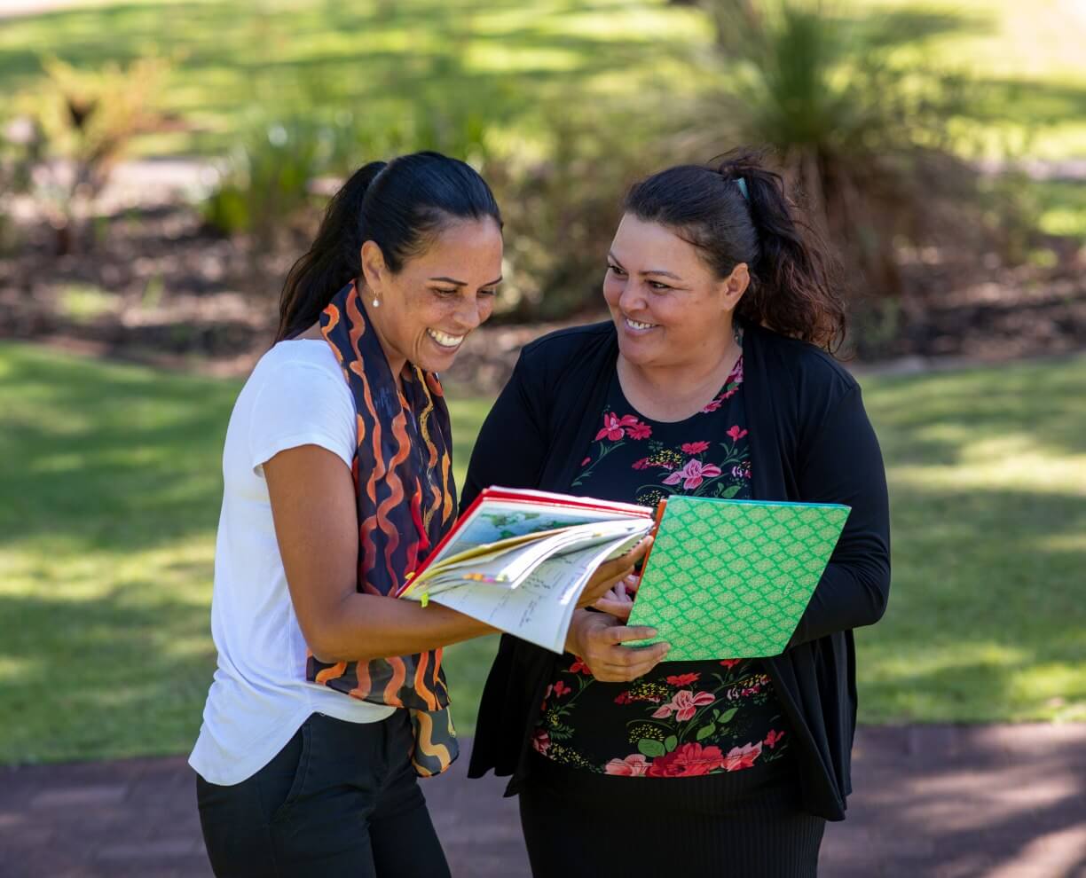 Two women standing outside holding books and smiling at each toher