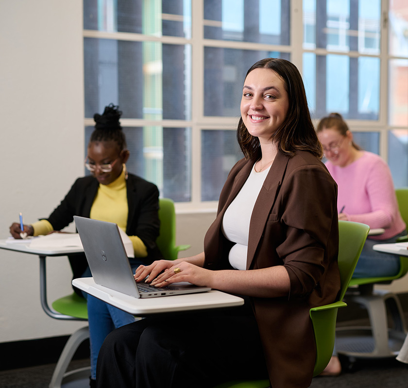 ACAP-Adelaide-classroom-840x800 A female student working on a laptop in a classroom and smiling to camera, with two more female students working at desks in the background