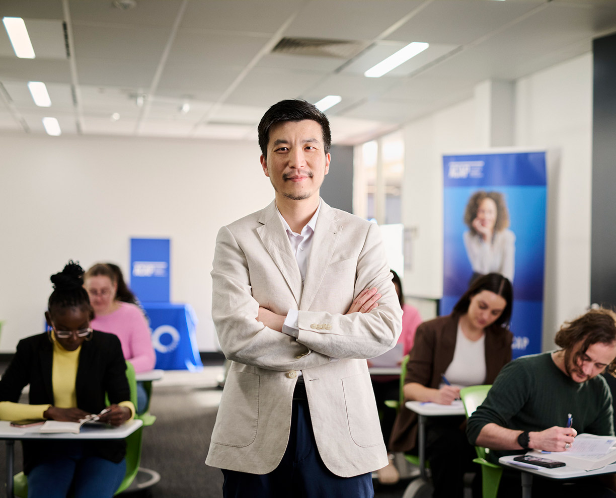 ACAP-career-outcomes-1224x990 A man with short hair wearing a beige suit jacket, standing with his arms folded in a classroom with ACAP University College students - career outcomes (1224x990)