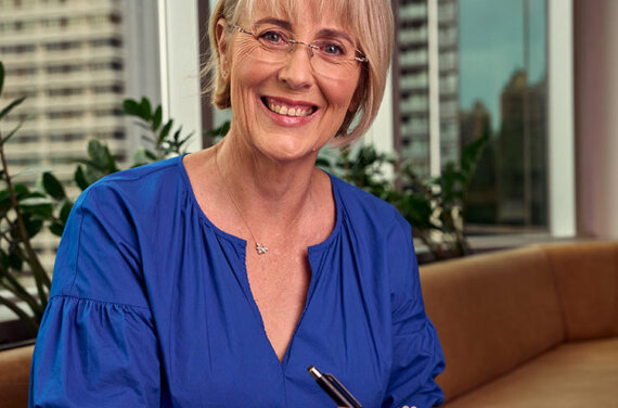 A female business coach sitting at a desk and writing in a book in front of a window with large office buildings in the background