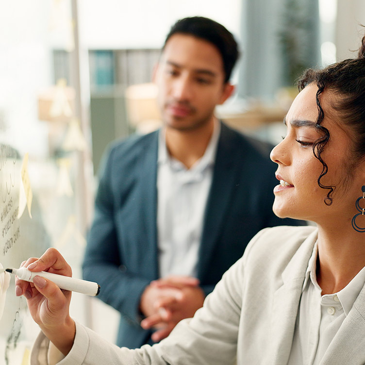 A female criminologist writing on a whiteboard with a marker pen in front of man in a suit watching in the background