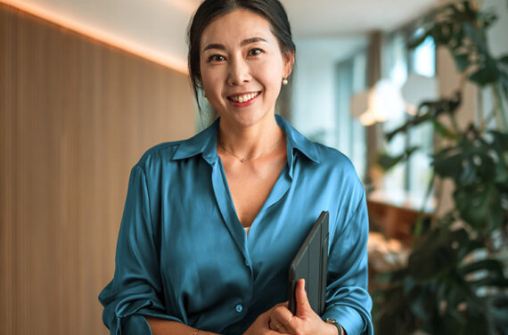 A woman standing in an office wearing a blue silk shirt and holding a tablet under her arm, smiling towards camera