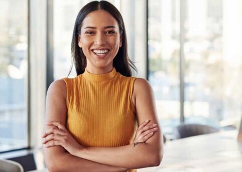 Woman stands with arms crossed. Broadly smiling. Glass windows and office in background