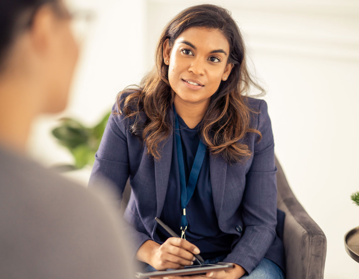 ACAP Woman sits in a chari, pen poised above clipboard. She wears a suit and a blue lanyard. Person blurred in foreground. They seem to be in a conversation.