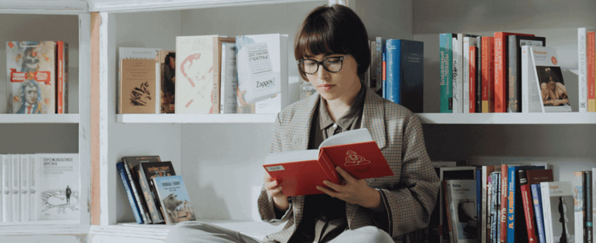 Woman sits cross legged on the floor in library. Books on shelf behind them.