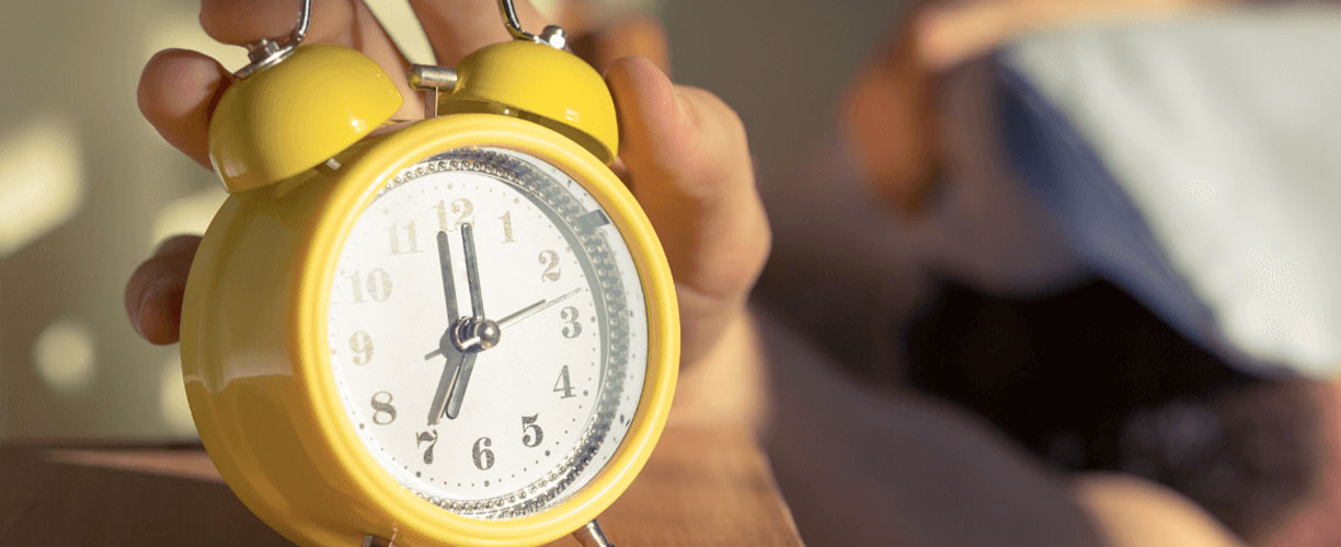 A yellow metal clock with two alarm bells atop reads 7. Bright light and shadows are seen on wall behind clock. A persons hand is resting on top of alarm bells.