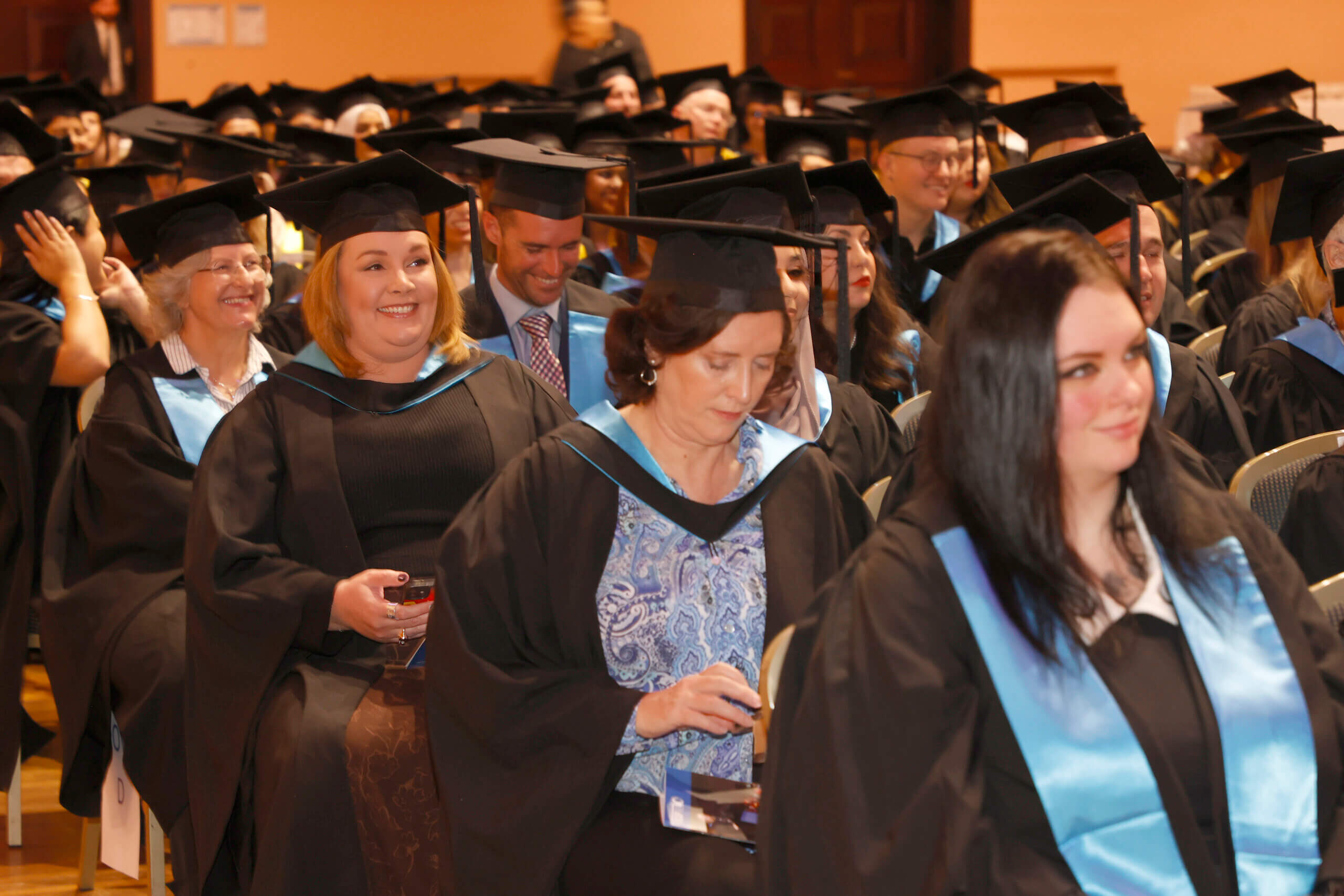 Graduation ceremony at ACAP Graduates sitting in hall/large space in ceremonial gowns and caps.