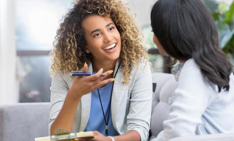 Presenter Person speaks to another person on a couch. She holds a pen and a clipboard and has her hand up.