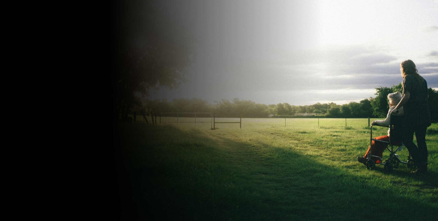Good Death Woman stands behind another person in a wheelchair. They are paused looking out at the distance in a green grass field. A fence is in the background.
