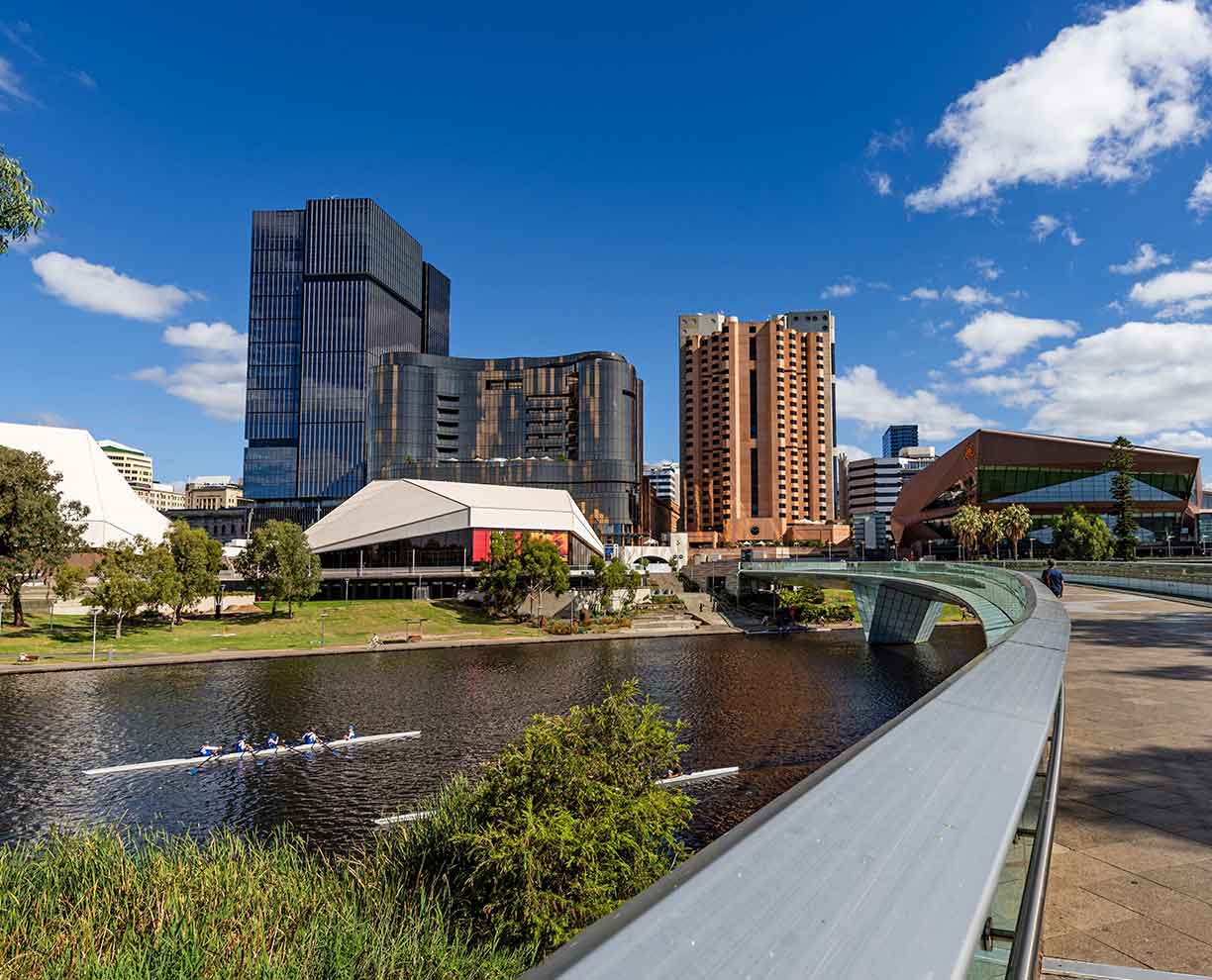 Adelaide Picture of outdoor cityscape. Pedestrian bridge crosses river. 4 person row boat on water. Blue skies and city buildings in the background. Adelaide in South Australia