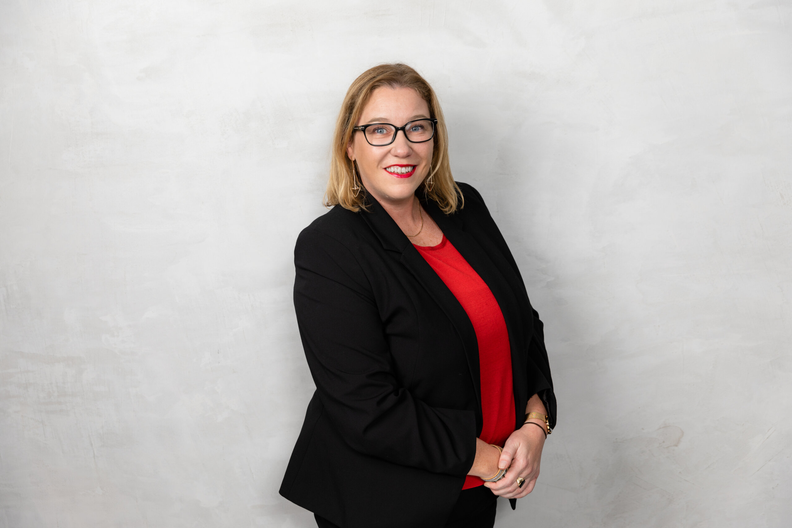 Kellie Hamilton Woman with blonde hair and glasses in corporate attire smiles in front of grey background