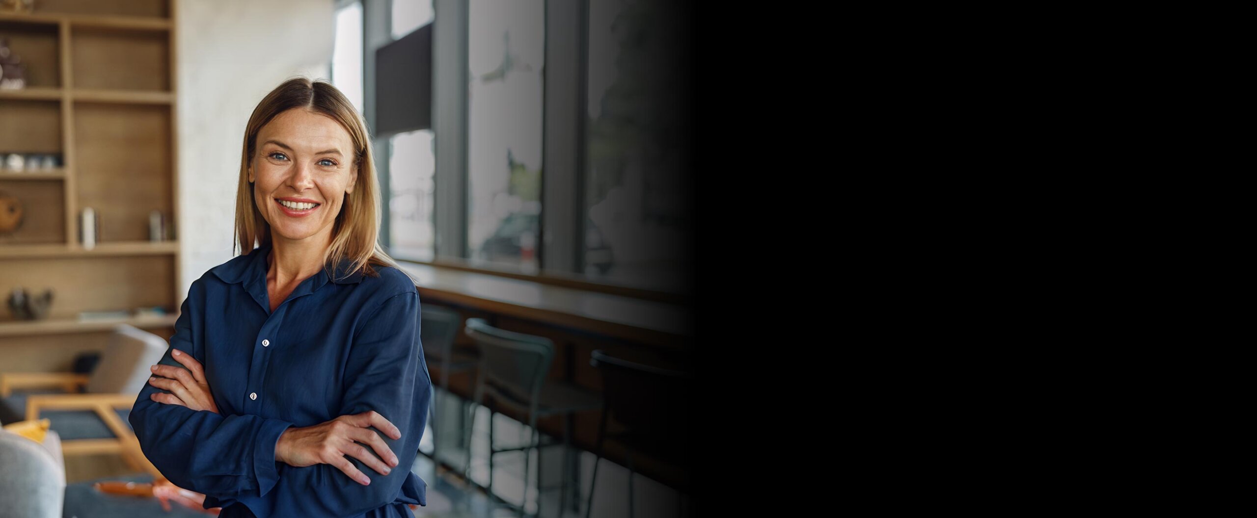A blonde-haired woman wearing a blue collared shirt, standing with her arms folded in front of a window
