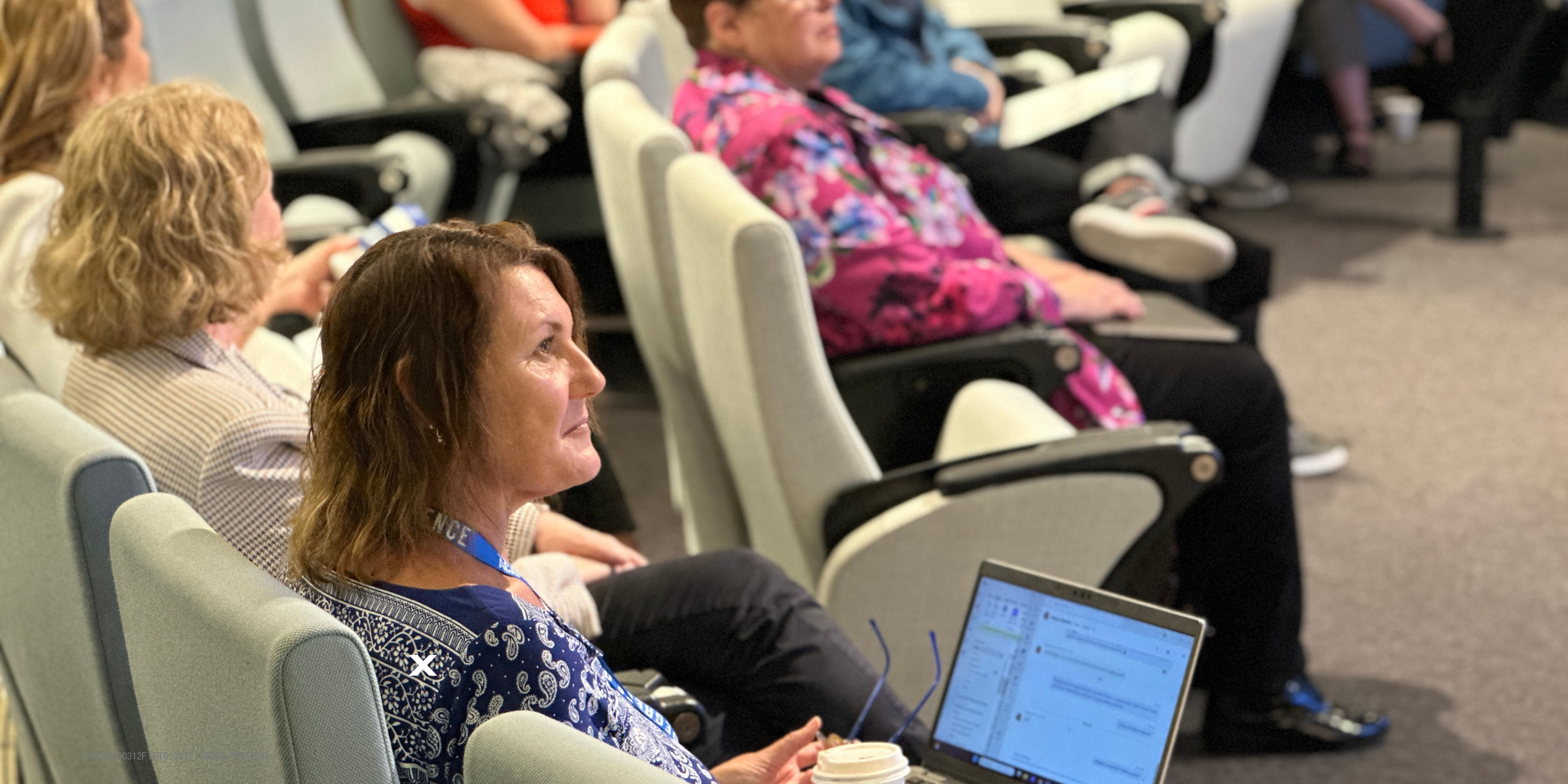 woman sits in lecture theatre filled with people. she has a coffee next to her. Laptop on her lap. And has removed her reading glasses.