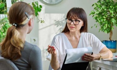 psychology career woman in office setting - sitting at desk with laptop on table. She holds a clipboard and pen in hand while speaking to a client | psychology career. A young person is seen in foreground, back to camera sitting in chair facing speaker