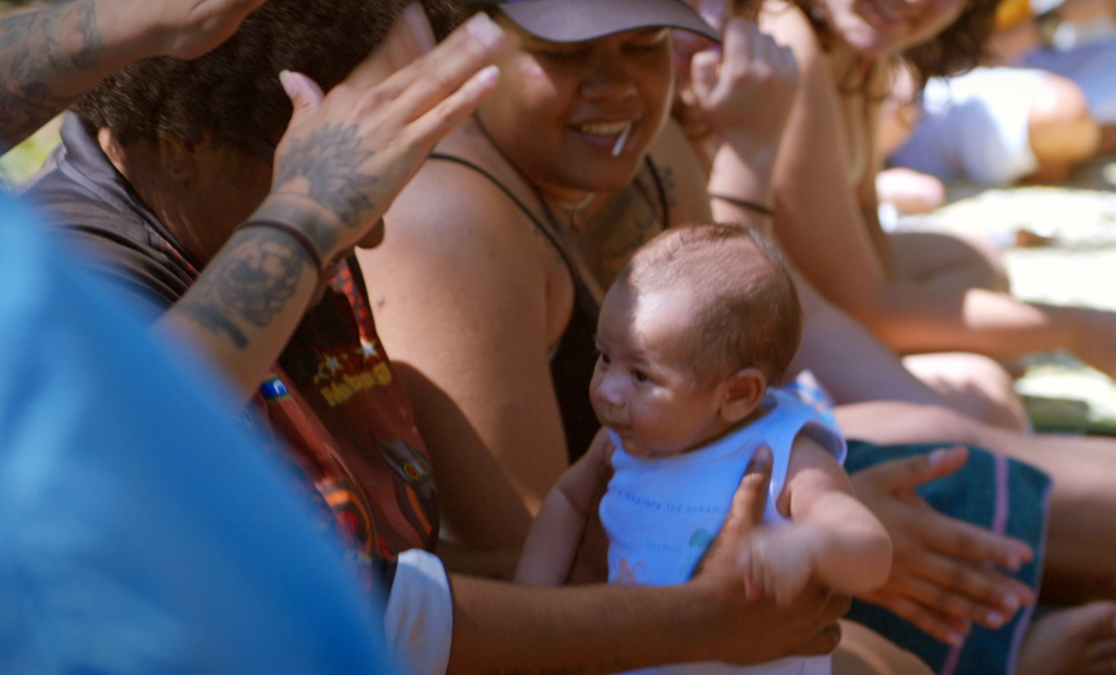 Krunal doco Image of baby being held in a group of people as someone raises tattooed hands above its heads