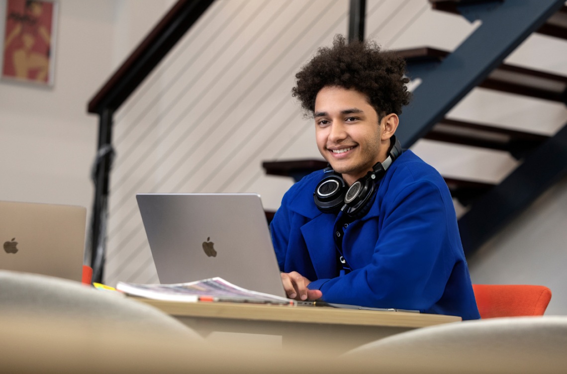 Criminology tile high school students A student with a blue jacket sitting at a desk, with a laptop open and headphones around his neck