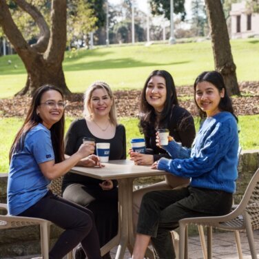 Four female high school students sitting at an outdoor table on campus with coffees in their hands - 761x761