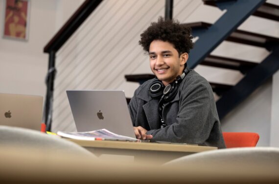 A student with a grey jacket sitting at a desk, with a laptop open and headphones around his neck