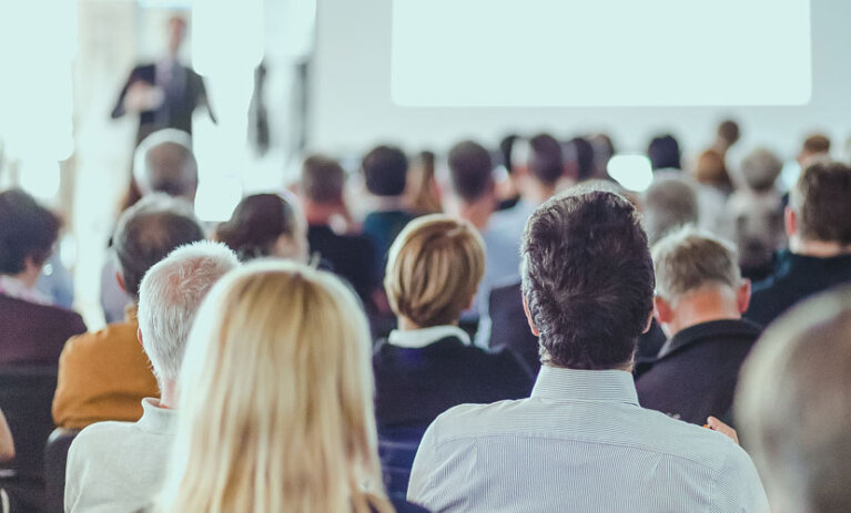 A group of academics sitting in chairs at a conference, facing with their backs to camera - ACAP Conference 828x676