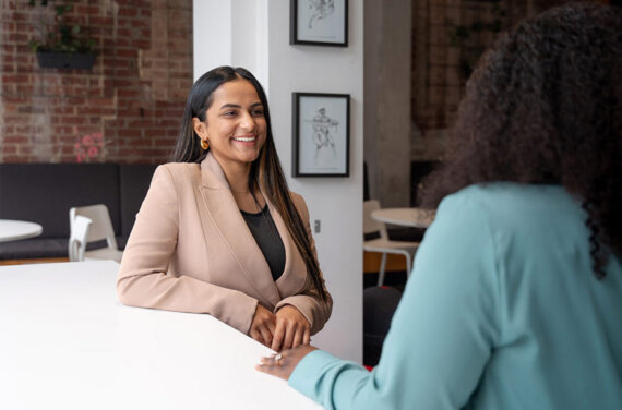 A female management student wearing a light pink suit jacket, sitting at a desk and smiling towards an ACAP lecturer in the foreground (1140x7532)