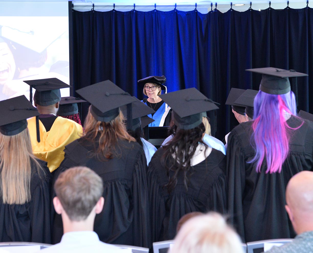 An ACAP lecturer standing on stage behind a lectern, with students in academic regalia standing in the crowd (1224x990)