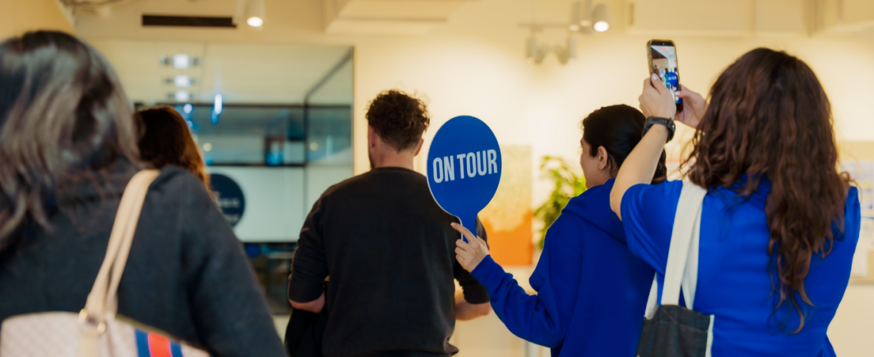 A group of visitors explore ACAP University College during a campus tour, led by a staff member holding a blue ‘On Tour’ sign, while another attendee takes photos on her phone.