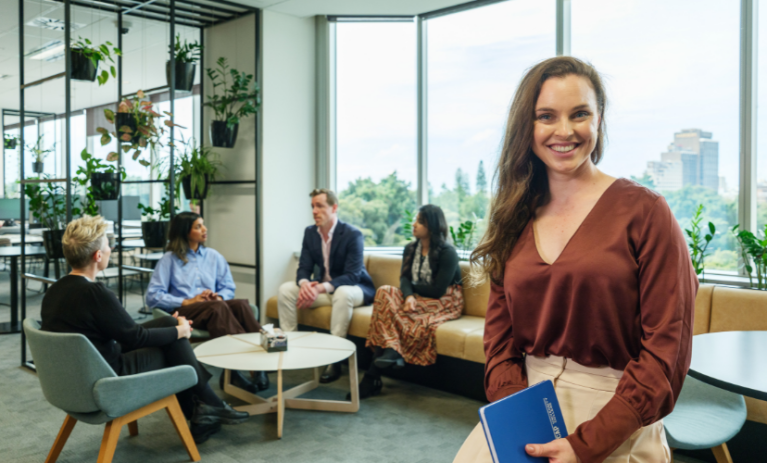 A smiling woman holding a blue notebook sits in the foreground of a bright, modern ACAP University College campus lounge, while four people chat in a seating area behind her.