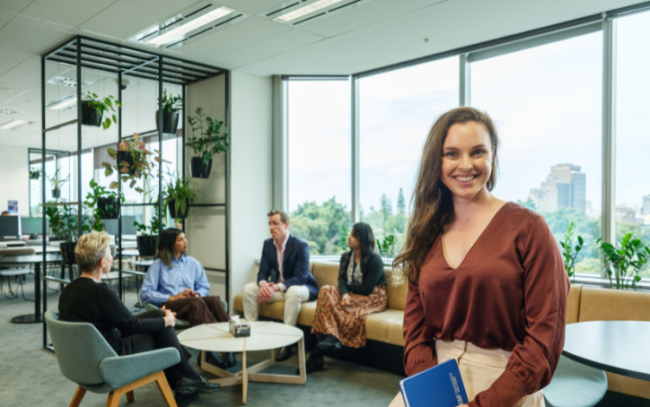 A smiling woman holding a blue notebook sits in the foreground of a bright, modern ACAP University College campus lounge, while four people chat in a seating area behind her.