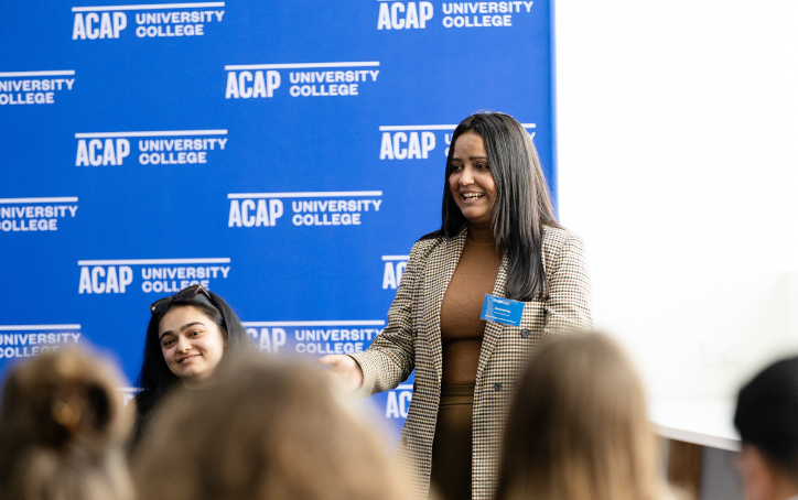 A presenter speaks to attendees at ACAP University College’s Melbourne Open Day, standing in front of a blue ACAP branded backdrop.