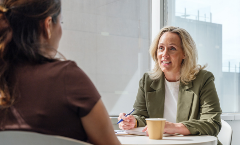 A smiling woman in a green blazer holds a pen while speaking with another woman across a table in a bright ACAP University College meeting space.