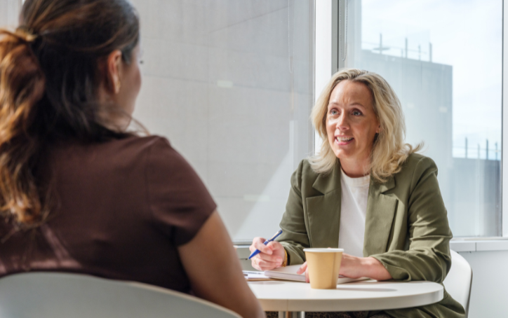 A smiling woman in a green blazer holds a pen while speaking with another woman across a table in a bright ACAP University College meeting space.