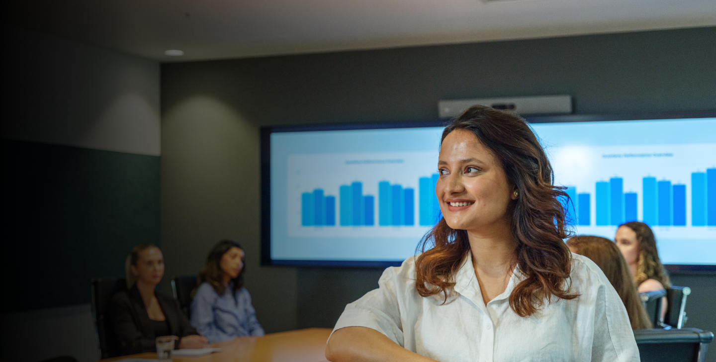 A smiling woman stands in the foreground of a modern ACAP University College meeting room, while five people sit around a conference table with presentation charts displayed on screens behind them.