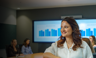 A smiling woman stands in the foreground of a modern ACAP University College meeting room, while five people sit around a conference table with presentation charts displayed on screens behind them.