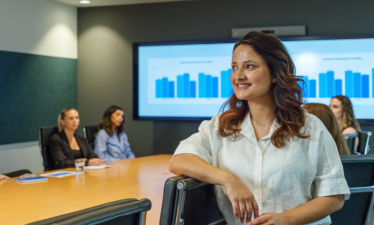 A smiling woman stands in the foreground of a modern ACAP University College meeting room, while five people sit around a conference table with presentation charts displayed on screens behind them.