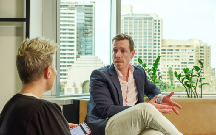 A man in a navy blazer gestures while speaking with another person in a bright ACAP University College lounge, with large windows overlooking the city.