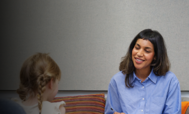 A smiling woman in a blue shirt sits on a couch holding a clipboard and speaking with a girl in a comfortable, modern counselling space at ACAP University College.