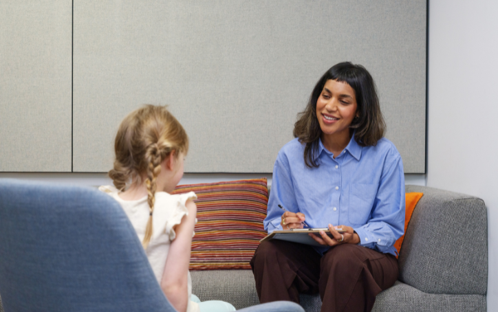 A smiling woman in a blue shirt sits on a couch holding a clipboard and speaking with a girl in a comfortable, modern counselling space at ACAP University College.